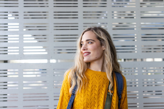 Woman standing in front of grey metal panel wall wearing mustard yellow sweater with backpack