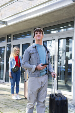 Diverse travel companions standing at modern entrance holding smartphone, rolling suitcases