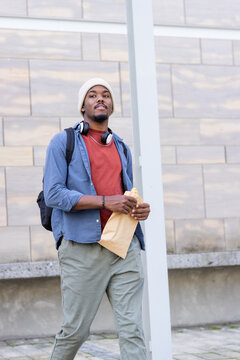 African american man holding envelope standing on column-lined walkway, backpack and headphones