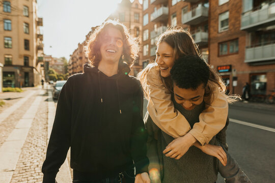 Portrait of smiling young man walking with male friend piggybacking female at street on sunny day