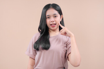 Asian woman in casual outfit smiling while pointing her finger at cheek, showing playful and confident expression against pastel background.