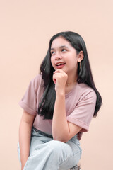 Asian woman with long black hair in casual modern outfit, wearing pastel t-shirt and denim, sitting with hand on chin, smiling, showing charm, elegance, and youthful confidence.