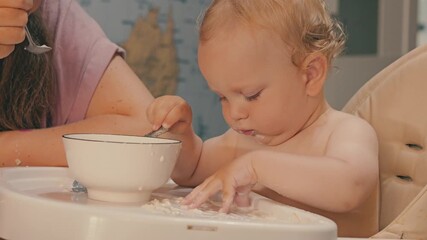 The kid is sitting at a high chair for eating. The parent feeds him porridge with a spoon. The child reaches into the bowl with his hand and scatters porridge on table. Close-up of baby's porridge - Powered by Adobe