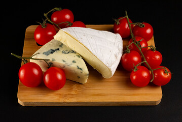 A delicious arrangement of gourmet cheeses, including brie and blue cheese, served with fresh cherry tomatoes on a wooden cutting board. Isolated on a black background.