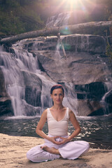 Young woman sitting in lotus pose under tropical waterfall meditating with calm strength and mindfulness fully connected to nature energy tranquility and inner balance