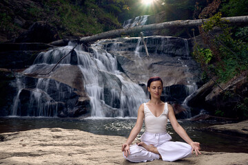 Young woman sitting in lotus pose under tropical waterfall meditating with calm strength and mindfulness fully connected to nature energy tranquility and inner balance