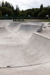 A spacious skatepark featuring smooth concrete ramps awaits skaters. The area is surrounded by greenery, with an overcast sky creating a calm atmosphere in the early evening