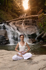 Young woman sitting in lotus pose under tropical waterfall meditating with calm strength and mindfulness fully connected to nature energy tranquility and inner balance