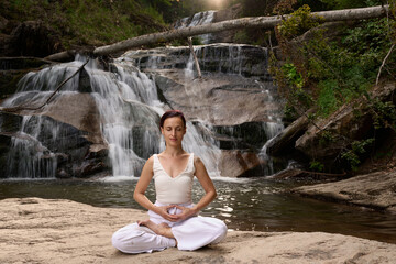 Young woman sitting in lotus pose under tropical waterfall meditating with calm strength and mindfulness fully connected to nature energy tranquility and inner balance