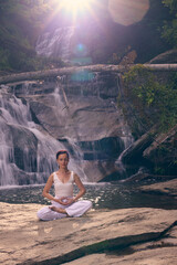 Young woman sitting in lotus pose under tropical waterfall meditating with calm strength and mindfulness fully connected to nature energy tranquility and inner balance