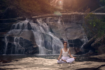 Young woman sitting in lotus pose under tropical waterfall meditating with calm strength and mindfulness fully connected to nature energy tranquility and inner balance