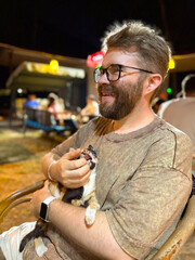 Man smiling while holding kitten in evening outdoor cafe. Human animal bond, companionship, and...
