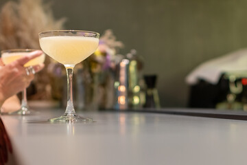 Perfectly crafted light yellow cocktail in a coupe glass on bar with another drink and person's hand blurred in background