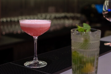 Vibrant pink cocktail with light foam top is served in coupe glass, placed on black bar mat next to mojito. The background is dark and out of focus