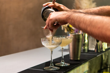 Bartender is expertly straining light-colored cocktail from shaker into two elegant coupe glasses....