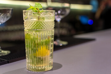 Refreshing cocktail in textured glass on bar, garnished with fresh mint leaves and citrus wedge. The background is dark and out of focus