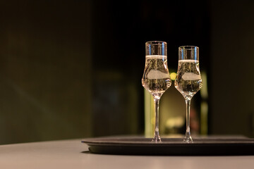 Two glasses filled with a clear, sparkling liquid sit side-by-side on a round, black tray. The background is dark and blurred, with a warm light reflecting off the glasses