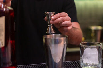 Bartender in black shirt is holding jigger over metal shaker, preparing to pour. A bottle of red liquid is on the left, and glass with ice is on the right