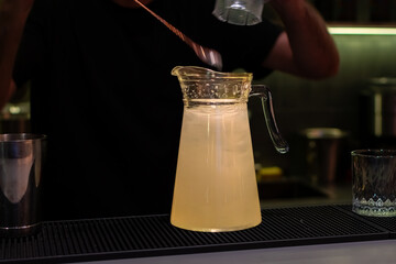 Large glass pitcher filled with pale yellow liquid and ice on bar mat on background of bartender in black t-shirt
