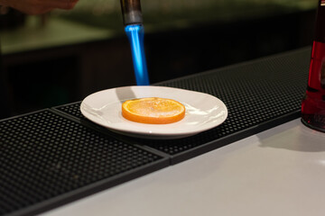 Slice of orange on small white plate is being torched with blue flame. The plate rests on a black rubber bar mat, with a bottle visible to the right