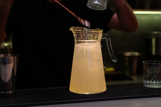 Large glass pitcher filled with pale yellow liquid and ice on bar mat on background of bartender in black t-shirt