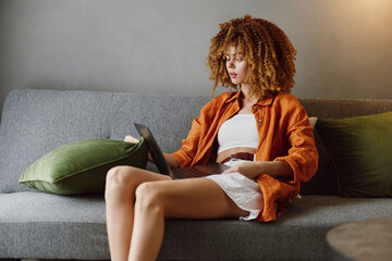 Young woman sitting on a sofa, focused on her laptop, wearing an orange shirt and white shorts Cozy...