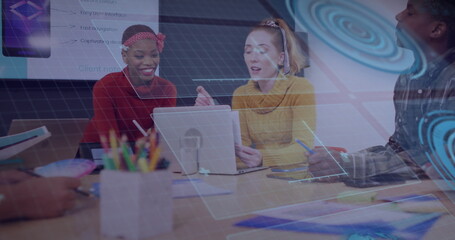 Colleagues in colorful sweaters discussing mockups at meeting table with laptop tablet and stylus