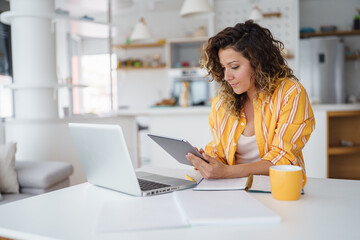 Professional Woman In Casual Yellow Striped Shirt Using Digital Devices While Working Remotely In Modern Bright Kitchen