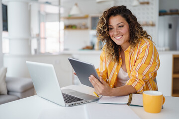 Fototapeta premium Young Woman Using Digital Devices For Remote Work While Sitting At White Table In Bright Modern Kitchen Space