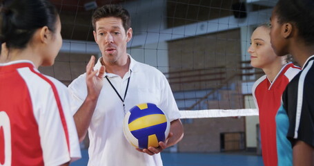 Male coach holding volleyball and signaling players on gym court, with net, jerseys and court lines