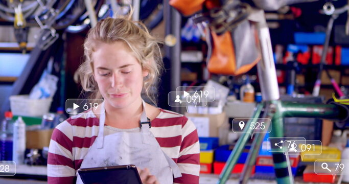 Inspecting mechanic in apron using tablet in bike shop with hanging wheels tools and green frame