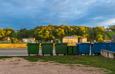 Row of green and blue recycling containers set against a backdrop of sunlit trees and small town...