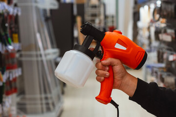 Hand holding bright orange electric paint spray gun with white container in hardware store aisle....