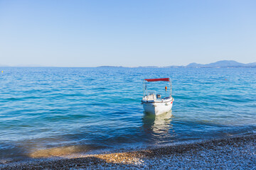 A small white motorboat with a red canopy is anchored in the clear shallow water just off a pebble beach, with a tranquil sea and a distant coastline.
