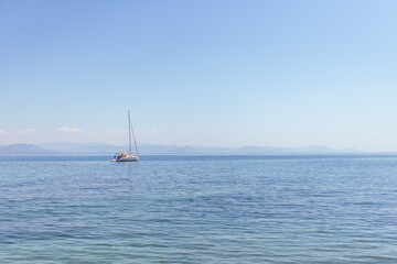 Naklejka premium A solitary sailboat floats peacefully on a vast, tranquil open sea, with a serene horizon line and distant mountains visible under a clear, bright blue sky.