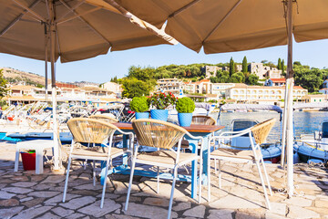 A tranquil outdoor cafe terrace with tables and chairs, offering a panoramic view of a sunny Mediterranean marina with boats and a historic village with a fortress.