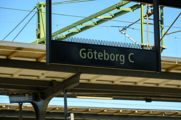 A train station sign displays the name Göteborg C under a bright sky. The surrounding structure and overhead wires suggest bustling regional transport activities