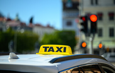 Yellow taxi cab sits at a red traffic light in a bustling city. Sunlight highlights the details of the vehicle and surroundings while pedestrians fill the streets nearby