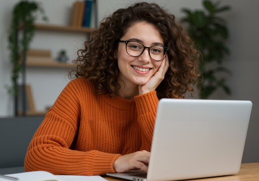Happy young woman with glasses working from home on her laptop, smiling at the camera in a modern interior
