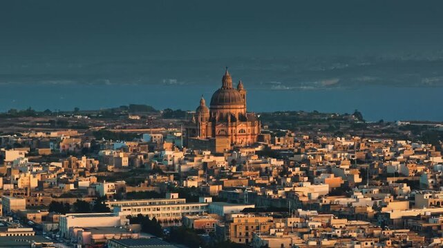 Malta, Comino island: Warm sunset light illuminating the Parish Church of the Assumption of the Blessed Virgin Mary or Xewkija Rotunda. City buildings and sea under dramatic sky. Drone flight footage