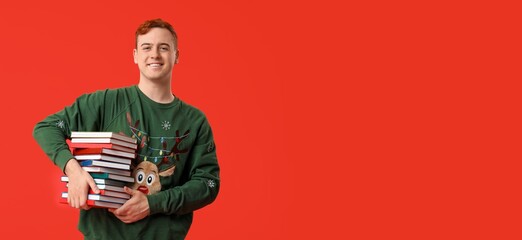 Happy young man in Christmas sweater holding many books on red background with space for text