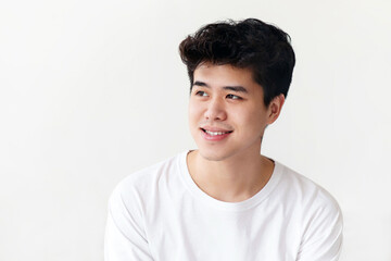 Modern teen at home at free time or weekend. Cheerful asian zoomer or young male in white t-shirt sits on bed, rest and enjoys good morning in light bedroom interior and looks towards empty space