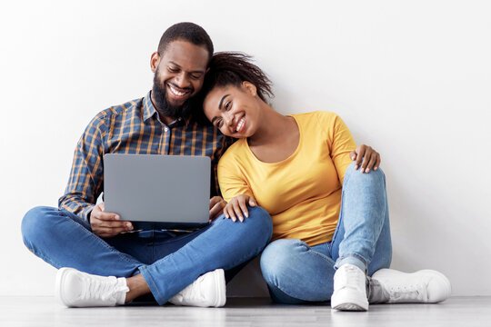 Cheerful young black man and woman look in laptop, planning interior, sits on floor in empty room on white wall background. Relocation, buy furniture for house or working on design together, panorama