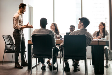 Corporate Meeting Concept. Young Smiling Businessman Giving Speech During Meeting With Coworkers In Modern Office, Standing At Desk In Board Room, Explaining Strategy And Management