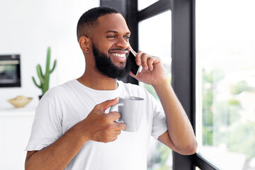 Pleasant Conversation. Portrait of smiling African American man talking on mobile phone in the morning standing near window holding cup of hot coffee, making call with friends or family