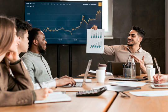 Smiling employer showing paper with graph diagram presentation, screen monitor with chart in the background at office, diverse workers listening to presentation. Financial analysis, business meeting