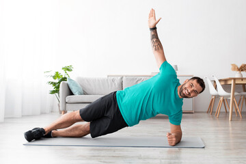 Happy young muscular caucasian man athlete in uniform doing side plank on floor in living room interior. Sport at home, workout and new normal, fitness and exercises, active lifestyle, empty space