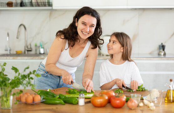 Delighted mother and little girl are engaged in cutting ingredients for salad on kitchen-table
