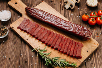 Sliced cured sausage with whole piece on wooden board and spices, rustic food styling