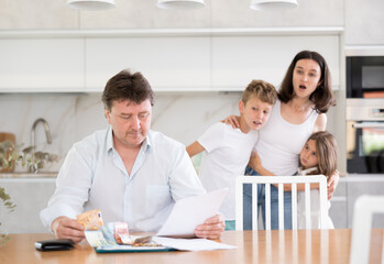 Sad father looks at bills and counts money. Upset wife with children in the background